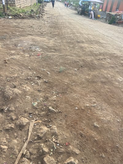 The image displays an unpaved dirt road in Gitothua ward, Kenya, viewed from a low-angle perspective. The foreground features an uneven, compacted earth surface strewn with rocks, small stones, and scattered debris, including plastic remnants and organic material. Visible tire tracks indicate vehicular passage. Along the left margin of the road, a low stone wall is visible, accompanied by a loose pile of larger stones and sparse vegetation. Further along this side, a blue sedan is parked. Several individuals are present on the road: one person in dark attire walks away from the viewer, and other figures are distributed further into the background. On the right side of the road, multiple makeshift market stalls are active. One stall includes a light-colored canopy positioned over a utility vehicle or display area, where an individual is bending over, interacting with merchandise. Adjacent to it, another stall, constructed from corrugated metal sheeting, showcases produce on a wooden table. The scene depicts a functional, active commercial thoroughfare with pedestrian and vehicular movement.