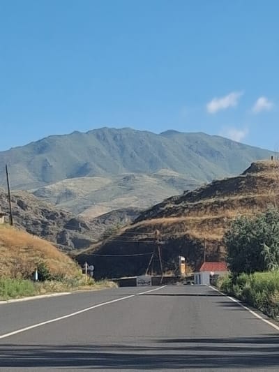 The image captures a scenic view of a mountainous landscape under a clear blue sky. A paved road with white lane markings leads towards a distant, majestic mountain range covered in green vegetation. The terrain on either side of the road is rugged and dry, with rocky outcrops and sparse vegetation.
On the right side of the road, a small building with a red-tiled roof is visible, nestled amongst greenery. There are a few signs and utility poles scattered along the roadside, indicating human presence and infrastructure. The overall impression is one of a rural, natural environment, possibly a remote area or a road passing through a mountainous region.
While the image depicts a generic mountainous scene and a road, it does not contain any specific visual cues that would allow for a definitive identification of the location as Yeghegnadzor, Armenia. To confirm the location, additional information or landmarks would be required.