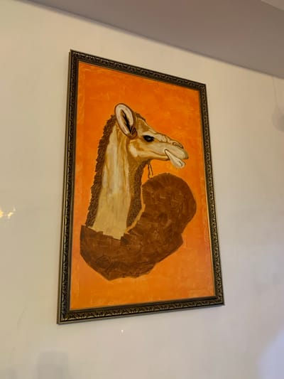 A framed painting on a white wall features an abstract depiction of a camel against an orange background. The camel's head is in profile, with a prominent eye and an open mouth revealing teeth. A large, brown shape, composed of angular segments, resembles a wing or a section of the camel's body. The painting has a textured appearance, possibly due to brushstrokes. A decorative, dark frame borders the artwork.