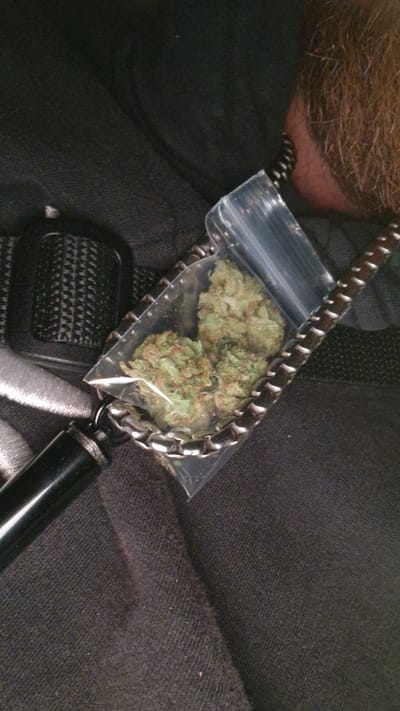 A close-up photograph shows a small, clear plastic bag containing what appears to be cannabis buds. The bag rests on a metal chain, a seatbelt buckle, and a black pen-like object. Part of a person's head with short, brown hair is visible in the upper left corner of the frame. The background consists of dark-colored fabric. There is no information to place this image within the city of Paralimni, Cyprus.