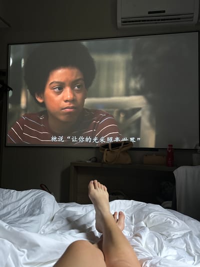 The image depicts an indoor scene, likely a bedroom, in Yangpu District, China. A large projection screen displays a close-up of a young Black boy with an afro, wearing a striped shirt. Chinese subtitles are visible at the bottom of the screen. In the foreground, a person's legs and feet are visible, resting on a white duvet. A brown tote bag and a red bottle are placed on a shelf behind the screen. An air conditioning unit is mounted on the wall above the screen. The lighting suggests a dimly lit room, with the projection screen being the primary light source.