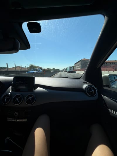 The image captures a view from inside a car, looking out through the windshield and side window. The car's interior is visible, including the dashboard with air vents and a central infotainment screen displaying track information for "Automobile For The Prophecy" by R.E.M. The perspective suggests the car is in motion on a highway.

Outside, the scene is dominated by a clear blue sky, indicating a sunny day, likely midday given the bright sunlight. There are buildings with tiled roofs visible along the side of the road, suggesting an urban or suburban environment. A truck with a green logo is seen in traffic ahead. The presence of other vehicles indicates that a normal traffic flow is occurring.

On the right side of the interior, a person's legs are visible, illuminated by sunlight casting a shadow. The person appears to be a passenger. The overall mood is one of travel on a pleasant day.

Visible text includes "AIRBAG" on the dashboard, "UnipolMoveR" on a device mounted on the windshield, and the song title and artist on the infotainment screen. The location context provided places the scene in Castellanza, Italy.