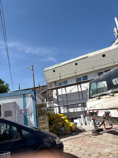 The image shows a construction site in Saedinenie, Bulgaria. A white building with scaffolding is visible. There's a blue car parked near the construction site. Rolls of yellow insulation material (ISOVER) are placed in a wire cage. A truck with the license plate "PA 4034 BY" is present. A white portable office unit with the text "Cтрои" and "www.stroymontazh.com" is also visible. A CCTV camera is mounted on a pole. The sky is blue with wispy clouds.