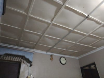 The image shows an interior view of a room, likely in a residential setting. The ceiling features a grid of beige panels, with white trim outlining the grid. On the wall, a round clock with a white face and black numbers is visible. To the left of the clock, a decorative hanging object, possibly a chandelier or an ornament, is suspended from the ceiling. In the lower left of the image, a portion of a doorway or large framed object is visible, with intricate dark trim along its top edge. The walls are a neutral, light color. The overall impression is of a simple, modestly furnished room. The image is not geographically specific to Iganmu, Nigeria, and no inhabitants or actions are depicted.