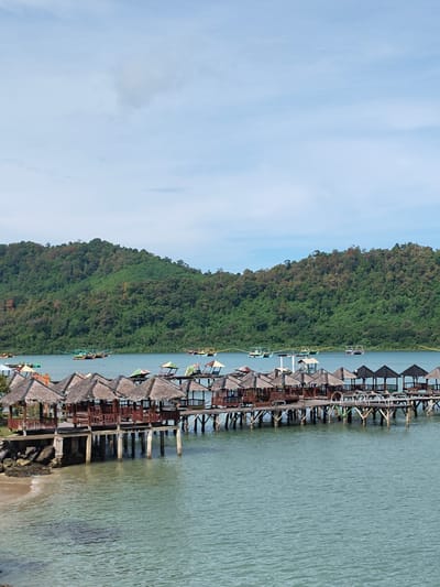 The image depicts a waterfront scene in Layeun, Indonesia. A long pier with huts extends into the water. The huts have thatched roofs and are supported by wooden pillars. In the background, there's a forested hill and several boats on the water. The sky is mostly blue with a few clouds. The water is a light blue-green color.