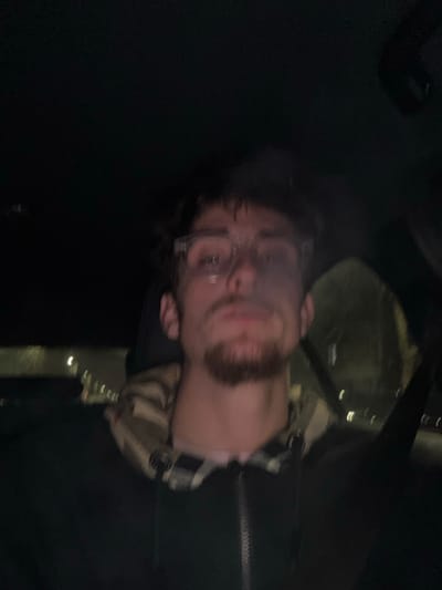 A blurry image shows a person with glasses and a beard looking at the camera, rotated ninety degrees counterclockwise. They appear to be wearing a cap and a checkered shirt. The background is dark, with hints of what may be a car interior or city lights visible. The overall image quality is poor.