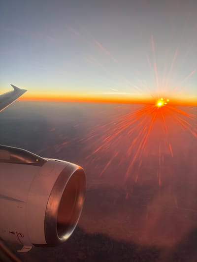 An aerial view from an airplane window captures a breathtaking sunrise over the landscape of Şarkışla, Turkey. The sun, a brilliant orb of light, is positioned on the horizon, casting long, fiery rays that streak across the hazy atmosphere. The sky transitions from a pale blue at the top to a vibrant orange and yellow near the horizon. The wing of the airplane extends from the left side of the frame, with the engine housing prominently displayed in the foreground. The landscape below is obscured by a layer of clouds or atmospheric haze, painted in deep reds and oranges by the sunrise.