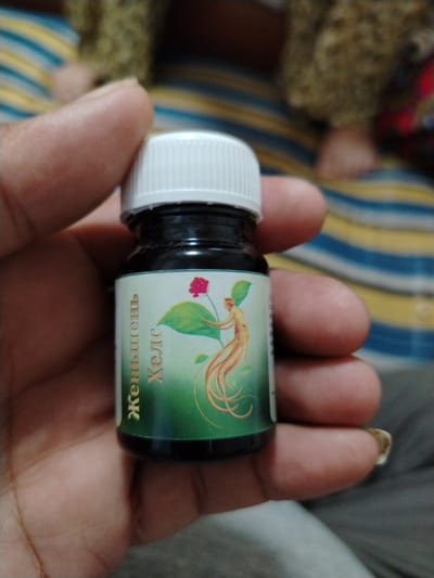 A person's hand holds a small, dark glass bottle with a white cap. The bottle has a label with a green background. On the label, there is a stylized illustration of a ginseng root and plant. The text on the label is in Cyrillic and appears to be "Женьшень" and "Хелс". The background of the image is blurred, showing striped fabric in yellow, blue, and white, and a person's leg. The lighting suggests it is daytime. The overall scene appears to be casual, possibly indoors.
