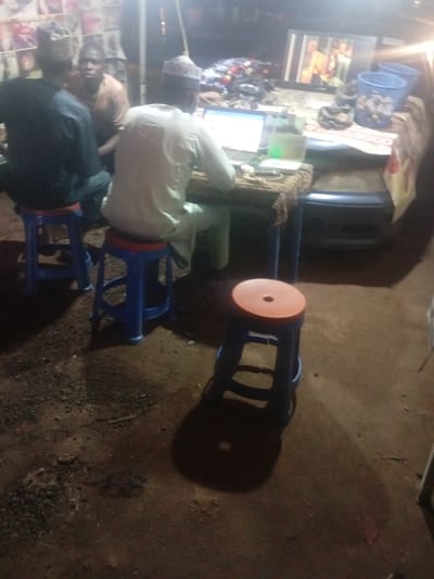 In the city of Tundun Wada West, Nigeria, this nighttime scene depicts an outdoor setting with a table laden with items, including a laptop and a television monitor. Three individuals are present, with two seated on blue stools and one standing. The lighting appears to be artificial, possibly from a nearby lamp. The ground is uneven and appears to be dirt with scattered small stones. In the background, a vehicle is partially visible. The general ambiance suggests a casual, communal gathering or a small business operation.