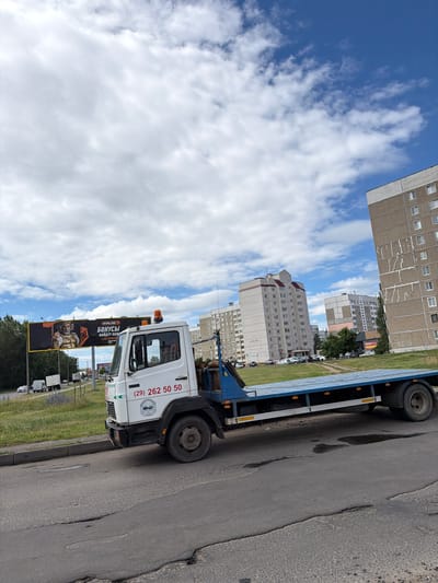 This media file captures a daytime scene in Mahilyow, Belarus, under a partly cloudy sky with the sun shining. The perspective is from a low angle, looking upwards and sideways, as if the camera itself is tilted.
The main subject is a white tow truck, parked on the side of a road. The truck has a blue flatbed trailer attached. Visible on the side of the truck's cab is the text "(29) 262 50 50". There is also a black billboard visible behind the truck, with Russian text that appears to be an advertisement. The billboard features an image of a man in a helmet and reads "WINLINE" and "БОНУСЫ НАЙДУТ КАЖ!".
In the background, several multi-story apartment buildings are prominent, characteristic of urban Soviet-era architecture. These buildings are a light gray or beige color and have rows of windows. The road itself is asphalt and shows signs of wear, with some potholes. To the left of the road, there is a strip of green grass and a line of parked vehicles, mostly white trucks. The overall atmosphere suggests an ordinary day in a residential urban area.