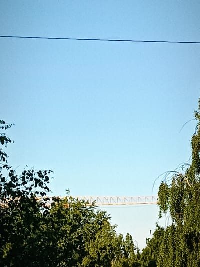 The image shows a bright blue sky with a thin dark horizontal line across the upper portion. The lower half of the image is filled with lush green foliage from trees. In the middle distance, peeking through the trees, is a section of a yellow construction crane or bridge with a distinct triangular pattern. The scene is likely an outdoor environment. There is no indication of the city of Arkhangelsk, Russia.