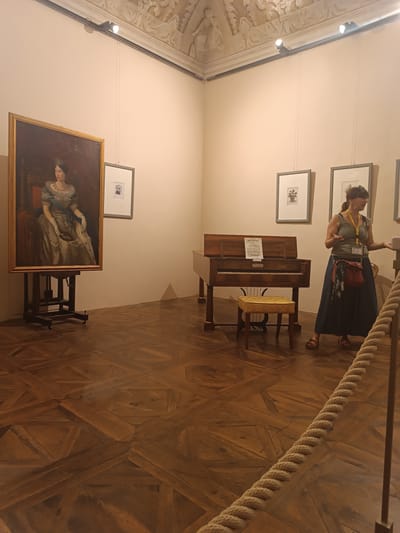 In a museum gallery in Colorno, Italy, a woman stands near a historic piano, gesturing as if explaining something. She is wearing a green tank top, a long navy skirt, and sandals. A red purse hangs from her shoulder, and she has a lanyard with badges around her neck. The room features a richly patterned wooden floor and a high ceiling with ornate plasterwork.
To the left of the piano, a large framed portrait of a woman in a formal gown and crown is displayed on an easel. Next to the portrait, two framed prints hang on the wall. The piano itself is an antique instrument with sheet music on its stand and a small yellow stool in front of it. The lighting in the room is provided by spotlights mounted on the ceiling. The scene appears to be set during the daytime, as indicated by the ambient light and artificial lighting. The overall atmosphere suggests a guided tour or educational presentation within the museum.
