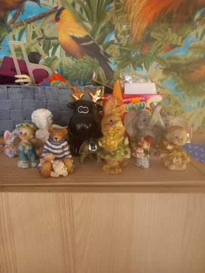 The image displays a collection of small decorative figurines and toys arranged vertically. The subjects include a black figurine of a moose with golden antlers and a white number '8' on its head, a teddy bear wrapped in a blue and white striped blanket, and a few smaller teddy bear figurines. There is also a figurine of a blonde-haired child wearing a green wreath and blue clothing, and another figurine of a white child. These are placed on a textured blue fabric surface. Behind the figurines, a vibrant fabric with a tropical bird and foliage pattern is visible. A pink plastic clothes hanger is also partially visible. To the right, the edge of a light-colored wooden surface is present. A pink and yellow package with Cyrillic text is visible amongst the items. The text "ТОЛЬКОРЫ" and "ВЖНЯ" and "5.00" are visible on the package. The overall impression is a collection of whimsical decorative items. The lighting suggests it is daytime.
