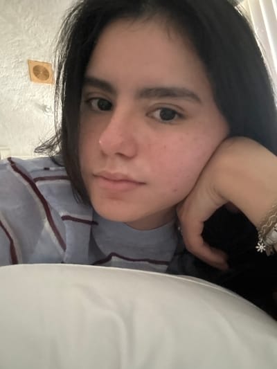 The image presents a close-up, waist-up indoor portrait of a young individual with dark hair, dark eyes, and light skin displaying visible freckles on the nose and cheeks. The individual gazes directly forward towards the camera with a neutral facial expression. They are attired in a light blue long-sleeve garment featuring horizontal maroon stripes. The individual's left hand is positioned against the left side of their face, supporting the chin area. Multiple bracelets, including chain-link designs and one featuring a white daisy-like charm, are visible on the left wrist. The foreground is partially occupied by a white pillow or bedding material. In the background, a white or off-white wall with a visible stucco or textured finish is present. Mounted on the upper left portion of this wall is a light-colored, rectangular object with an internal circular, possibly spiral or fan-blade design. The visual information within the image does not provide any identifiable markers for a specific location within Santiago, Mexico.