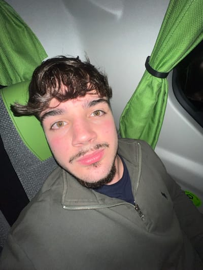 A young man with hazel eyes and dark, wavy hair rests his head on a green pillow inside a vehicle. He has a beard and mustache. He wears a dark blue undershirt and a zipped-up olive green jacket, revealing the neck of the shirt. A green curtain is tied above him, near the vehicle's window. The interior wall is white, and the seat fabric is grey.