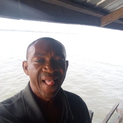 An adult male with dark skin and short dark hair is depicted in a close-up, chest-up view, situated in Koko, Nigeria. He faces the camera, extending his tongue in a playful gesture. Perspiration is visible on his forehead. He wears a dark, textured collared shirt. Above the subject, a wooden overhead structure, including beams and a panel, is present. The background features a broad expanse of light brown/grey, rippled water, consistent with a river or bay, extending to a faint landmass on the distant horizon under a bright, diffused sky. A wooden plank structure is partially visible to the subject's right, extending towards the water.