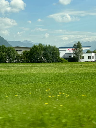 The image depicts a daytime exterior scene in Bruck an der Großglocknerstraße, Austria. In the foreground, a blurred expanse of bright green grass stretches across the frame, dotted with small yellow wildflowers. The middle ground features a line of dense green trees separating the field from a white industrial building with a blue roof. The building has several windows and a sign with red lettering that reads "ATTENSCH..." on the right side. To the left of this building, another white structure with a similar roofline is visible, also with green trees in front of it, and the partial text "ofrost*" can be seen on its wall. In the background, a range of hazy, blue-toned mountains forms the horizon beneath a sky with scattered white clouds. The overall impression is a rural or semi-industrial setting with a focus on a natural landscape.