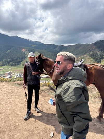 This outdoor image, taken in Jeti-Ögüz, Kyrgyzstan, is rotated 90 degrees clockwise, depicting a mountainous landscape under a cloudy sky.
In the foreground, a white man with blonde hair, a beard, and sunglasses is captured mid-laugh or shout, his mouth wide open in an expression of clear joy. He wears a dark olive green jacket and holds a crumpled blue paper with faint white text in his left hand. The head of a brown horse is positioned close to his face, suggesting interaction, possibly feeding or playful engagement.
Standing behind and to the left of the main subject is another person, appearing to be an Asian male. He wears a white cap with the word "VINTAGE" clearly visible, a dark sleeveless puffer vest over a burgundy long-sleeved shirt, and dark pants. He holds a stick or whip in his right hand and is positioned next to another brown horse. His expression is neutral.
The ground beneath the subjects is dry, dusty earth. The background showcases vast, tree-covered mountains that define the rugged terrain. A valley with a small village, characterized by scattered buildings with light-colored roofs, is visible nestled among the hills. The sky is overcast with a mix of grey clouds, indicating daytime conditions without direct sunlight. The scene suggests a pleasant horse riding excursion or tour in this scenic, rural mountainous region.
