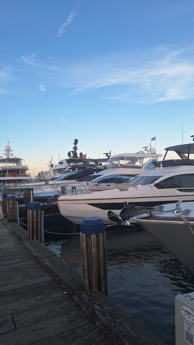The image depicts a row of large, modern yachts docked along a wooden pier. The yachts are sleek and white with dark accents and multiple levels. They are moored side-by-side, suggesting a marina setting. The water is dark and reflects the sky and the boats. The sky is a clear blue with some wispy white clouds, indicating it is daytime, likely late afternoon or early evening given the warm tones in the sky. The pier is made of weathered wood, with some pilings visible, each capped with a blue protective bumper.
On one of the yachts, a white rectangular device with the text "onSpot WiFi" and a QR code is visible. The number "21" is also painted on the side of a structure on one of the yachts. The scene appears peaceful and serene. The overall impression is one of luxury and leisure, typical of a high-end marina. The location is identified as Nantucket, United States of America.