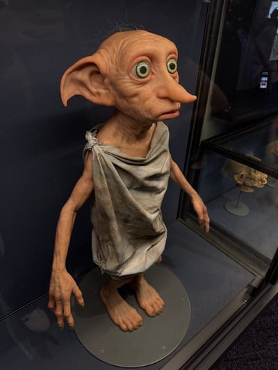 A life-sized figure resembling Dobby from the Harry Potter franchise is lying inside a glass display case. The figure is dressed in a grey cloth and has large, green eyes and pointed ears. The figure rests on a grey, circular platform. The scene is within a museum or exhibit, likely located in the city of Nerima, Japan. Other display cases and objects are visible in the background.