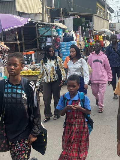 The image captures a busy daytime street scene in Egan, Nigeria, featuring multiple pedestrians and market stalls. The ground is a light gray paved surface.
On the left, a purple umbrella marked "MONEY TRANSFER" shades a market stall displaying household goods, including plastic basins, packaged items, and an appliance box. A mannequin torso in a leopard print top stands near this stall. Further right, more market stalls line the street, stocked with various items such as cleaning supplies, plastic containers, and other packaged products. Buildings with corrugated metal siding and overhead utility wires are visible in the background.
Six individuals are prominently featured, all walking generally from left to right across the frame.
1. In the immediate foreground on the left, a young person, likely female, wears a black and blue checkered long-sleeved shirt (resembling a Chelsea FC jersey) over floral-patterned pants. She carries a black backpack and a black bag with yellow patterns.
2. Behind and to the right, an adult female with dark braided hair wears a multi-patterned (gold, black, white) long-sleeved blouse and dark trousers. She carries a black shoulder bag and holds a dark beverage bottle and a smartphone.
3. Centered in the mid-ground, a young girl with short dark hair wears a blue short-sleeved top with a white collar over a long red plaid dress. She has a blue backpack and is seen manipulating a small, light-colored object in her hands while looking towards the viewer.
4. To her right, an adult female with straight dark hair wears a shiny white long-sleeved blouse and dark trousers, carrying a light-colored handbag.
5. Further back on the right, an adult male with short hair and eyeglasses wears a pink hooded sweatshirt with "EGAN UMMU" text and matching pink sweatpants. He wears sandals and carries a light-colored shoulder bag.
6. Partially visible on the far right, an adult male wears a dark blue collared shirt and dark trousers, carrying a dark shoulder bag.
The scene depicts active pedestrian movement within a commercial urban setting.