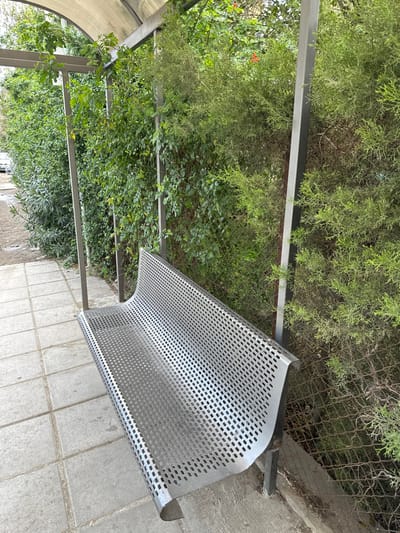 The image captures a quiet outdoor scene in Egkomi Lefkosias, Cyprus, featuring a modern, silver-colored metal bench with a distinctive perforated design. The bench, which has a gently curved shape, is set upon a paved ground consisting of light grey rectangular concrete tiles.

The setting is abundant with lush green vegetation. Behind and around the bench, dense foliage includes coniferous shrubs resembling cypress trees and various leafy plants, some of which appear to be growing over a metal framework or pergola structure. This structure comprises several horizontal metal beams, suggesting a shaded pathway or garden feature. A chain-link fence is partially visible through the dense greenery in the background, separating the landscaped area from a concrete curb or low wall.

The scene is tranquil and devoid of people or any ongoing activity. The bright, natural light indicates a daytime setting, likely under fair weather conditions, enhancing the vibrant green hues of the foliage. The combination of urban infrastructure like the bench and paving with natural, flourishing greenery suggests a public park, a garden pathway, or a well-maintained urban green space. No visible text is present in the image.