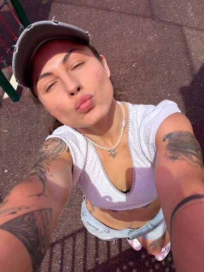A woman is lying on the ground, apparently on a playground surface, as inferred from the red rubber ground cover and colorful play equipment visible in the periphery. She is oriented at an angle towards the camera, suggesting she is taking a selfie. The woman wears a white top with a beaded necklace, denim shorts, and pink shoes. She has several tattoos on her arms and torso. She has her eyes closed and her lips pursed. The location is Kiryat Motzkin, Israel, based on available location data.