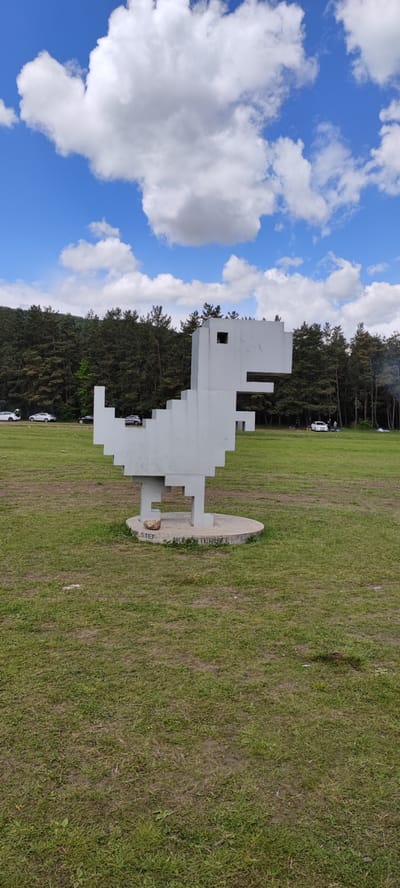 A full shot captures a large, gray, blocky dinosaur sculpture in a grassy field, against a backdrop of trees and a partly cloudy sky. The sculpture, resembling a retro 8-bit design, stands on a circular concrete base. The base has some graffiti/writing on it. In the background, several vehicles are parked, and people are scattered amongst the trees. The scene is located in Gyulagarak, Armenia.