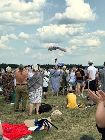 This image depicts an outdoor event likely taking place at an airfield or open field near Nestiukovo, Russia. In the sky, a parachute is visible, descending with a person attached. Below, a crowd of spectators stands on grassy ground, facing the sky and observing the parachute. The crowd consists of individuals of various ages and genders, dressed in casual attire suitable for warm weather, with some wearing hats and headscarves. Several people are holding hands or interacting with each other. Objects scattered on the ground include blankets, bags, and what appear to be picnic items. The environment is characterized by a large, open field under a cloudy sky, with a line of trees visible in the distance. The overall scene suggests a public event, possibly an airshow or festival, where skydiving is being demonstrated.