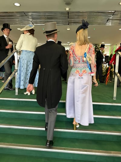The image captures an indoor scene at a multi-level facility in Epsom and Ewell, United Kingdom, likely during a formal event. The view is from behind, looking up a wide, green carpeted staircase featuring silver-colored metal nosings on each step.

In the immediate foreground, two individuals are ascending the stairs. A man on the left, seen from the back, wears a black tailcoat, light grey trousers, and black dress shoes, topped with a grey top hat. His right hand is slightly extended forward. To his right, a woman, also seen from the back, wears a long, light purple satin gown. The bodice and sleeves of her dress are adorned with intricate red floral and star embroidery, and it features an open back design. She wears white shoes with a gold block heel and carries a small, cream-colored shoulder bag with a gold clasp and a blue lanyard displaying an event pass. Her blonde hair is partially covered by a dark blue fascinator with a prominent bow. She holds a smartphone and another small item in her left hand.

Further up the stairs on the left, a man wearing a black top hat and dark suit is visible from the chest up, facing the viewer with a drink in a clear plastic cup in his right hand. Adjacent to him, a woman with her back mostly to the viewer wears a white bolero jacket over a light blue skirt featuring a lace-like floral pattern, and a wide-brimmed white hat with blue detailing.

In the background on the right, other formally dressed individuals are partially visible. One man wears a dark suit and top hat, and another figure in a red jacket holds a red and yellow striped umbrella. White floral arrangements serve as background decor. The ceiling above features recessed lighting and a reflective paneling system. Metal handrails are visible on the left side of the staircase.