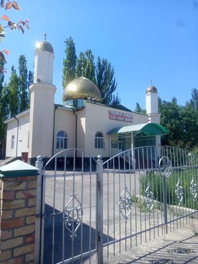 The image shows a mosque located in the city of Kara-Bulak, Kyrgyzstan. The mosque is a light-colored building with a prominent golden dome and a tall minaret. In front of the mosque, there is a metal gate with ornate decorations. The area appears to be well-maintained, with trees and greenery surrounding the building. The sky is clear and blue, indicating a sunny day. The overall scene conveys a sense of peace and tranquility.
