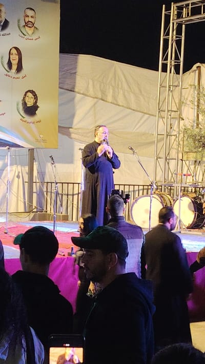 On a stage, a man in a black robe speaks into a microphone. Behind him, a backdrop displays portraits and names. Drums and scaffolding stand nearby. Several people watch from below, some wearing hats and dark clothing. A phone records the event. The setting appears to be an outdoor gathering, possibly at night.