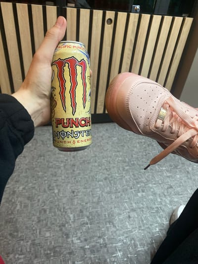 A person's left hand holds a yellow can of Monster Energy drink, labeled "PACIFIC PUNCH" and "PUNCH MONSTER". To the right of the can, a light pink sneaker is visible. The background consists of vertical wooden slats. The scene is indoors, likely on a floor with a grey patterned carpet. There is no indication of the location being Severodvinsk, Russia.