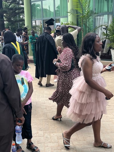 The image displays an outdoor gathering of approximately a dozen individuals on a paved area in Madina, Ghana, against the backdrop of a modern building with large green-tinted glass windows and surrounding tropical foliage. The ground is composed of light-colored interlocking pavers.
In the immediate foreground, a woman wearing a light pink, multi-tiered ruffled dress and silver flat sandals walks from right to left, holding a black smartphone in her left hand. To her left, a woman in a long-sleeved maroon dress with a white speckled pattern and dark heeled mules also walks left, displaying a watch on her left wrist and carrying a dark shoulder bag.
On the far left foreground, a young individual in a pink, blue, and yellow tie-dye t-shirt with a green badge pinned to it, black trousers, and black sandals stands looking downwards, partially obscured by a person in a brown long-sleeved shirt holding a 'PERLE' branded water bottle. Another 'PERLE' bottle is visible near their feet.
In the mid-ground, several individuals are present, notably some wearing black academic gowns. One person in a black gown and mortarboard is seen from behind, walking away from the camera. Another person in a black gown has a visible green and yellow academic stole. Further back, a person in a light tan t-shirt has their hand on their head, and another individual is seen filming with a smartphone. A man in a green outfit stands further in the background. Potted palm plants are visible near the building facade.
