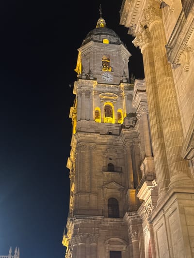 The image shows the bell tower of the Malaga Cathedral (Catedral de la Encarnación de Málaga) at night. The tower is illuminated with warm yellow lights, highlighting its intricate stone architecture, including decorative carvings, arched windows, and balconies. A clock face is visible on the tower. To the right of the tower, a large section of the cathedral's facade is visible, featuring massive fluted columns and ornate stonework. The sky is dark, indicating it is nighttime. The perspective is from a low angle, looking up at the imposing structure. The overall scene conveys a sense of grandeur and historical significance.