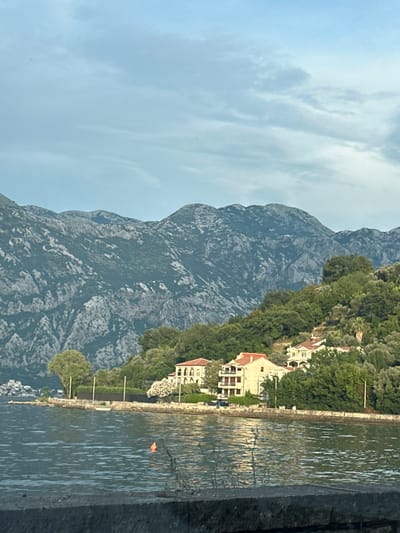 The image depicts a scenic view of the town of Prčanj, Montenegro, along the Bay of Kotor. In the foreground, a calm body of water, likely part of the bay, reflects the light from the sky. A small orange buoy is visible in the water. The middle ground features a coastline with several buildings situated on a lush, green hillside dotted with trees. The architecture of the buildings appears to be Mediterranean in style, with terracotta roofs. The hillside slopes upwards towards a majestic mountain range that dominates the background. The mountains are rugged and rocky, with sparse vegetation in places, and are partially covered by shadows. The sky is overcast with scattered clouds, suggesting a diffused natural light. The overall impression is one of natural beauty and tranquility.