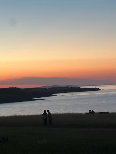 The image depicts a serene sunset over the Black Sea near Sinemorets, Bulgaria. Silhouetted against the vibrant orange and pink sky, two figures stand on a grassy hill overlooking the water. The sea is calm, reflecting the warm hues of the sunset. In the distance, the coastline is visible, with dark, rolling hills meeting the water. Further out, a faint line of mountains stretches across the horizon. The foreground is dominated by dark, silhouetted grass, with faint hints of other figures or objects present in the darkness. The overall atmosphere is peaceful and contemplative, capturing the beauty of a coastal sunset.