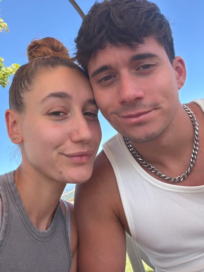 A close-up selfie features a young couple embracing outdoors, with a bright blue sky and hints of greenery in the background. The woman, on the left, has her hair in a bun and is wearing a grey tank top and small earrings. The man, on the right, has dark hair and is wearing a white tank top and a silver chain necklace. They are positioned very close together, with their heads tilted towards each other, suggesting an intimate moment. The environment appears to be sunny and warm, possibly during the daytime. The context of Kocherinovo, Bulgaria is not discernible from the image itself.