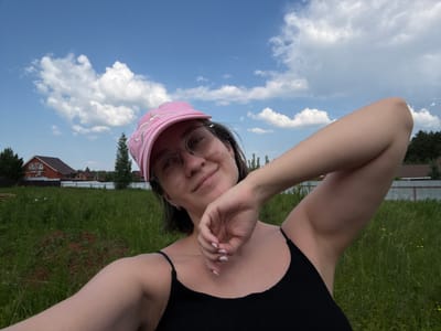 A selfie taken outdoors under a blue sky with white clouds shows a young woman wearing a pink baseball cap and glasses. She is looking towards the camera with a slight smile, her right arm raised and bent at the elbow. She is wearing a black tank top. In the background, there is a grassy field with scattered small yellow flowers, some trees, and houses with reddish-brown roofs. The scene appears to be in a rural or suburban area, possibly on the outskirts of a city or town, and is not specifically identifiable as being in the city of Dokshytsy, Russia based on visual cues alone.