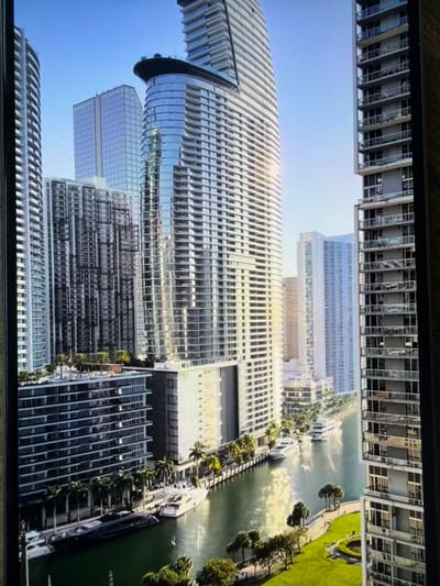 The image displays a modern urban waterfront scene, likely a digital rendering or a stylized photograph, viewed as if rotated 90 degrees clockwise from its natural orientation. When viewed upright, numerous contemporary high-rise buildings dominate the skyline, featuring extensive glass facades, balconies, and sleek designs. A prominent central skyscraper exhibits a distinctive, curved, and textured white exterior, brightly reflecting the sunlight.
These tall structures line a wide, greenish-blue body of water, resembling a river or canal. Several small boats or yachts are docked along the banks, suggesting recreational or transport activity. The waterfront is flanked by expansive green areas, indicative of parks or landscaped promenades, adorned with various trees, including palm-like species, which suggest a warm climate. Paved paths or roads run through these green spaces.
The scene is set under a clear, bright blue sky, indicating sunny weather with strong sunlight casting reflections and glares on the buildings' surfaces. There are no identifiable people or specific events clearly visible. The overall impression is a grand, developed metropolitan area. No visible text is present in the image. The visual characteristics of the scene, particularly the architecture and vegetation, do not correspond with the typical landscape of Razlog, Bulgaria.