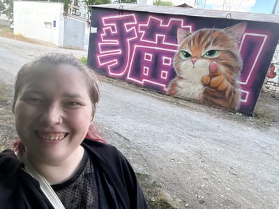 This media file shows a selfie taken outdoors, likely in Lipetsk, Russia. The photo features a young woman with light brown hair and pink streaks, smiling at the camera. She is wearing a black top with a sheer, dotted neckline, and a black jacket. Her face is the primary subject in the foreground, positioned upside down due to the selfie's orientation.

Behind the woman, a large, colorful mural is visible on a wall. The mural depicts a detailed, cartoonish orange cat with bright green eyes. The cat is painted in a realistic style, with visible fur texture and shading. To the right of the cat, there are stylized, abstract purple shapes outlined in white, possibly forming text or decorative elements. Some graffiti is also visible on a brick wall to the far left of the mural.

The background suggests an urban or industrial setting. The ground is rough concrete or asphalt, with patches of dry grass and dirt. In the distance, industrial buildings, including what appears to be a garage or warehouse with a metal door, are visible. The lighting suggests it is daytime, with bright sunlight casting shadows. The overall mood of the image is cheerful and casual, conveyed by the woman's smile. There is no visible text that can be clearly deciphered as a language.