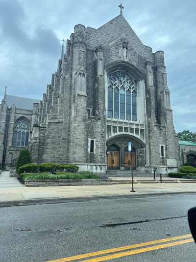The image shows the exterior of a large, ornate stone church, likely Gothic in style, located in Oak Park, Illinois, United States. The church is positioned close to the sidewalk and street, which has double yellow lines indicating a no-passing zone. The church features an arched entrance with large wooden doors and a large, elaborately decorated window above the entrance. The façade is constructed from gray stone blocks. There is landscaping along the sidewalk with various shrubs and flowering plants. The sky is overcast. The image is taken from a slightly low angle. A car side mirror is partially visible in the top right corner, indicating the photo was likely taken from a vehicle.