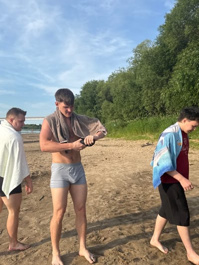 Three young men are standing on a sandy beach. The man in the center is wearing grey boxer briefs and has a tan towel draped over his shoulders. He is holding a black object in his hands, possibly a phone or a small device, and appears to be interacting with it. To his left, another man is wearing a white towel around himself and dark swim shorts. He is looking towards the man in the center. To the right, a third man is walking away from the camera, wearing a maroon t-shirt, black shorts, and a blue towel with a dolphin pattern draped over his shoulders. The background shows a body of water with a bridge and a small boat in the distance, and lush green trees line the edge of the beach. The sky is blue with scattered clouds. The location is the beach in Ryabinino, Russia.