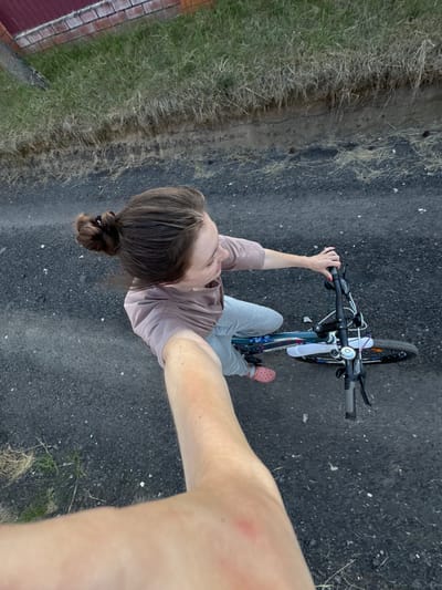 An overhead shot shows a young woman with her hair in a bun, wearing a light pink t-shirt and gray sweatpants. She is holding the handlebars of a bicycle, which is positioned in front of her. The bicycle has a white water bottle attached to the frame. She is wearing pink Crocs. Her right arm is extended towards the camera, obscuring part of the scene. The woman is standing on a dark, asphalt-like surface. To the left of the woman and bicycle, there is a patch of dry grass and dirt, with a brick wall in the background. The scene appears to be an outdoor setting, possibly on a road or path in the village of Krivandino, Russia.