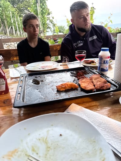 The image depicts a table outdoors with two individuals, likely at a restaurant or a private gathering in Dolenje Grčevje, Slovenia. In the foreground, a baking tray with remnants of cooked food, possibly chicken or pork, sits on a wooden table. A white plate with leftover food and cutlery is also visible. To the left, a bottle with a red cap, possibly ketchup, stands near a glass of red wine. A bottle of yogurt or milk with a blue and white label is present on the right, next to a roll of paper towels. The background shows a green, leafy vine-covered structure, suggesting a rural or vineyard setting. One person, a young male with short, dark hair, is seated on the left, wearing a black t-shirt. On the right, a man with short hair and a beard, wearing a dark purple hooded jacket, is seated. The lighting suggests it is daytime, possibly late afternoon or early evening.