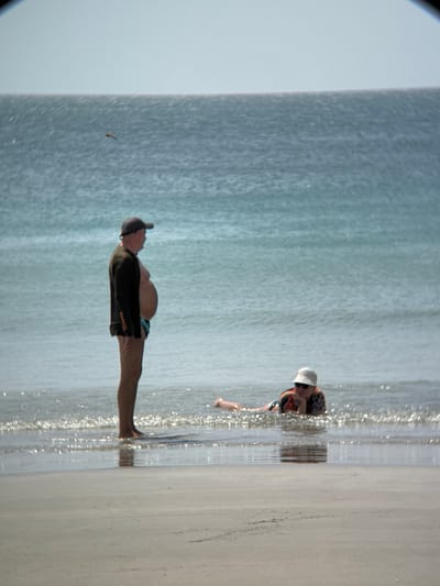A man stands in shallow water at a beach. He is wearing a dark long-sleeved shirt and dark shorts. He has a baseball cap on and is facing to the right. His stomach is protruding. In the background, a person lies in the water wearing a hat and sunglasses, resting their chin on their hands. The water is calm and blue, with a light-colored sandy beach in the foreground. The sky is a pale blue and hazy. The location is not identifiable as Dong Ninh Hoa, Vietnam based on the image alone.