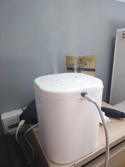 The image shows a white humidifier with a pink trim, emitting a faint mist. A grey USB cable is plugged into the humidifier, and another USB cable is connected to a black power bank, which is itself plugged into a white power strip. The humidifier and power bank are situated on a light-colored wooden surface, likely a nightstand or table. In the background, a beige pouch with a blue label reading "Seve" is visible against a grey wall. The scene appears to be indoors, possibly in a bedroom or living area, given the presence of a humidifier and the overall domestic setting. The lighting suggests it could be daytime, but the exact time of day is not discernible. There are no people visible in the image. The humidifier appears to be in operation, as indicated by the mist.
