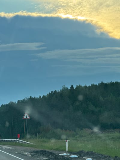 The image depicts a roadside scene in Karagay, Russia, under a cloudy sky with streaks of yellow sunlight illuminating the upper atmosphere. The foreground shows a paved road with white lane markings, bordered by a guardrail and grassy verges. Several small, light-colored puddles are visible on the road surface. A triangular road sign with a red border and a black cross symbol on a white background is prominently displayed on a post. To the left of the sign, a red rectangular object is partially visible. In the background, a dense forest of dark green coniferous trees stretches across the horizon, with a lighter, hazy layer of foliage closer to the road. The overall environment suggests a rural or semi-rural setting, likely during the daytime.