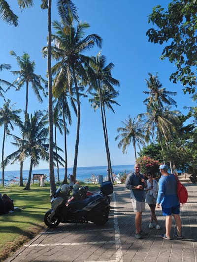 This image depicts a sunny day in Karangasem, Indonesia, likely near the coast. The foreground features a paved walkway with several parked scooters, one prominently in the center. Three individuals are visible on the walkway: a man in a blue shirt and grey shorts, a woman in a white top and denim shorts, and another man in a blue long-sleeved shirt and blue shorts, carrying a red backpack. They appear to be conversing. Behind them, a grassy area is dotted with palm trees, and the ocean with several boats can be seen in the background. The sky is a clear blue. To the far left, on the grass, are two individuals seated, facing away from the camera. The scene suggests a tropical tourist location.