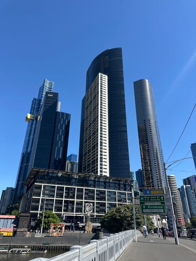 A street scene features modern architecture, including several high-rise buildings with reflective glass exteriors against a clear blue sky. Road signs provide directional information, pointing towards "Richmond" and routes to the "West Gate Br." A pedestrian bridge and walkway are visible with a metallic railing. The city skyline is complex, showcasing a blend of sleek, contemporary designs.